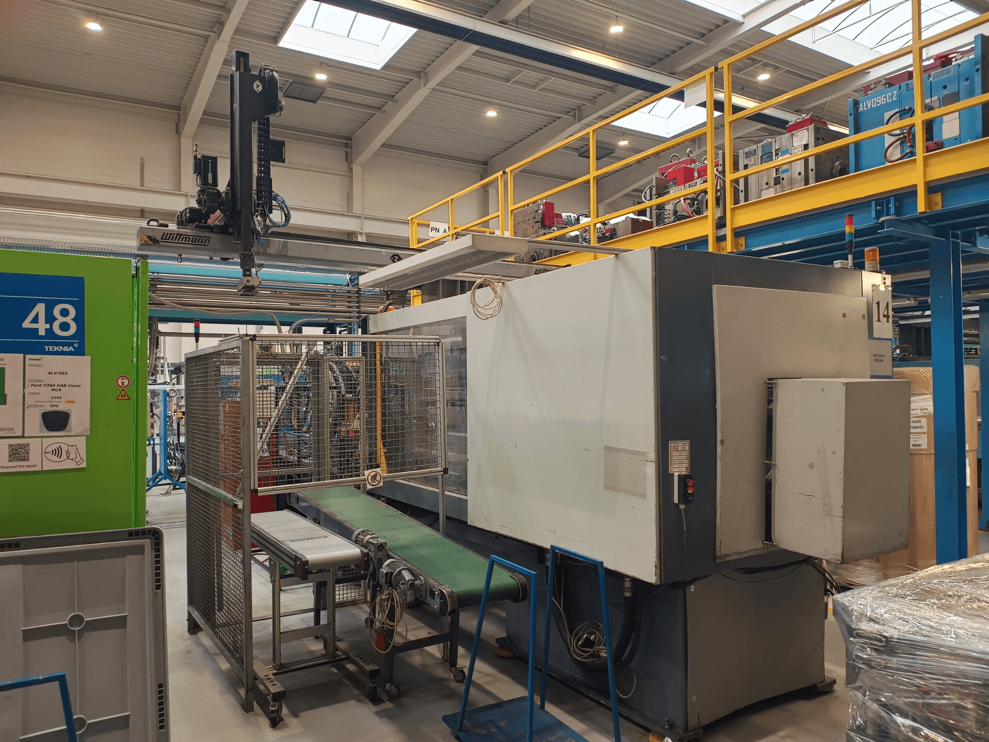 Urządzenia automatyki przemysłowej z zielonym przenośnikiem oznaczonym "48" i suwnicą; widok z boku maszyny na hali produkcyjnej.
